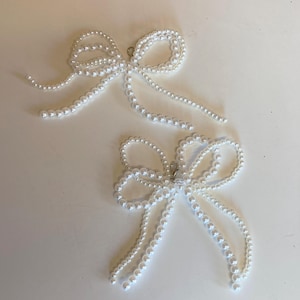 Puede incluir: Dos pinzas para el pelo de color blanco con forma de lazo de perlas. Cada pinza est&aacute; hecha de una cadena de perlas que est&aacute;n atadas en forma de lazo. Los lazos tienen colas largas que cuelgan.