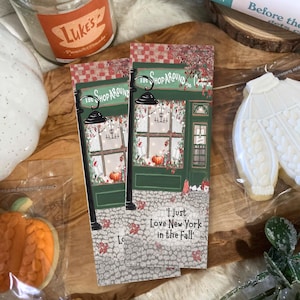 May include: Two rectangular bookmarks featuring a green storefront illustration with the text The Shop Around The Corner and I just Love New York in the Fall. The design includes a cobblestone street, a black lamppost, pumpkins, and autumn leaves.