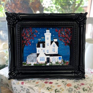 May include: A framed painting of a white house with a central tower against a dark blue night sky filled with stars. The scene includes two trees with red leaves, a small greenhouse structure, and a white picket fence surrounding the house.