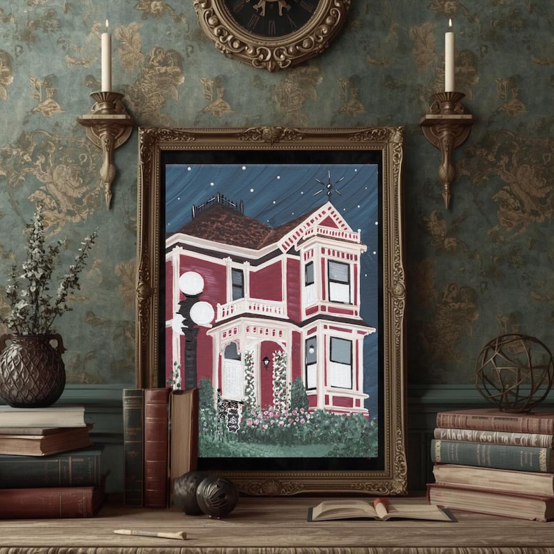 Puede incluir: Pintura enmarcada de una casa victoriana con exterior rojo, molduras blancas y un fondo de cielo nocturno azul oscuro. La obra de arte se exhibe sobre una superficie de madera con libros y objetos decorativos.