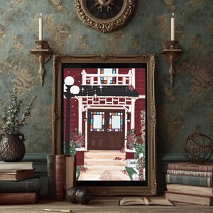 May include: A framed artwork depicts a red building with a white porch and stained glass windows. The artwork is in a gold frame and is displayed on a wooden surface with books, candles, and a decorative clock in the background.