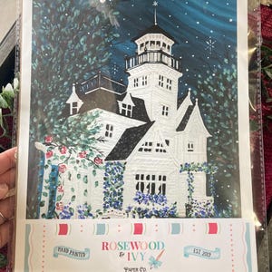 May include: Hand-painted art print of a white Victorian house with a tower, surrounded by trees and flowers. The artwork is set against a dark blue night sky with falling snow. The print includes the text "Rosewood & Ivy Paper Co. EST 2019."