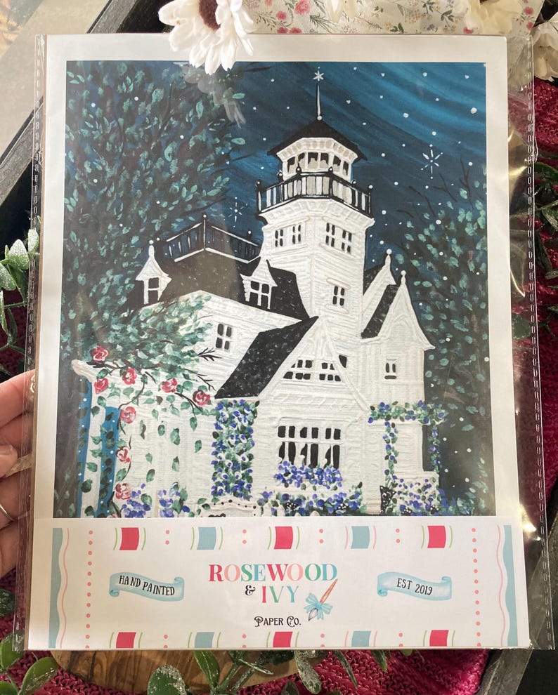 May include: Hand-painted art print of a white Victorian house with a tower, surrounded by trees and flowers. The artwork is set against a dark blue night sky with falling snow. The print includes the text "Rosewood & Ivy Paper Co. EST 2019."