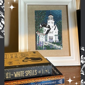 May include: A framed painting of a white house with a tower, surrounded by a white picket fence and flowers. The painting is in a white frame, and is set on a table with a stack of books, including one titled "Big Book of White Spells".