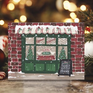K&ouml;nnte beinhalten: Weihnachtliche Illustration des Caf&eacute;s Central Perk mit roter Backsteinfassade, gr&uuml;nen Verzierungen und einem kleinen Tafelschild mit der Aufschrift "Happy Holidays Friends". Das Caf&eacute; hat eine festliche Markise und Weihnachtsb&auml;ume.
