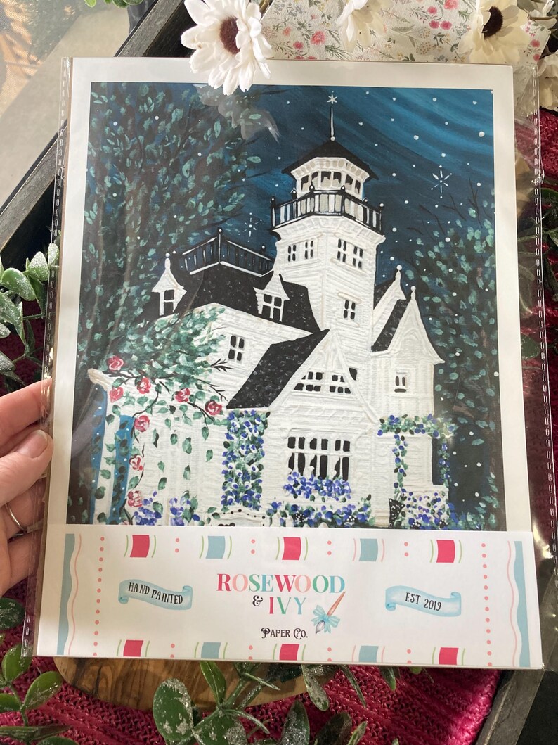 May include: A hand-painted art print of a white Victorian house with a tower, surrounded by lush greenery and blue flowers. The night sky is a deep blue with stars. The print is labeled "Rosewood & Ivy Paper Co."