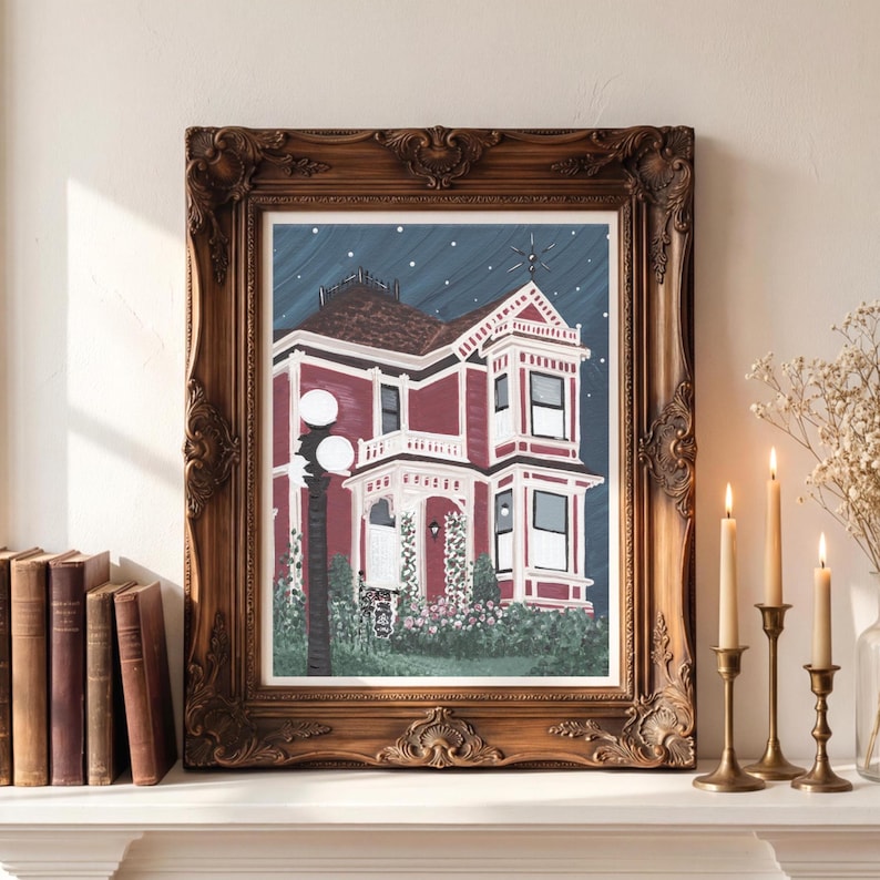 Puede incluir: Una pintura enmarcada de una casa victoriana con exterior rojo y adornos blancos, sobre un cielo estrellado. El marco ornamentado, de color marr&oacute;n oscuro, complementa la obra de arte. La pintura se exhibe en una repisa blanca, con velas y libros cerca.