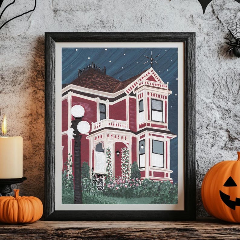 Puede incluir: Obra de arte enmarcada que representa una casa victoriana roja bajo un cielo estrellado. La pintura presenta una fachada detallada con molduras blancas, un tejado oscuro y vegetaci&oacute;n. Una vela encendida y una calabaza tallada sugieren un tema de Halloween.