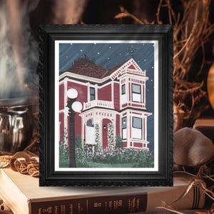 Puede incluir: Una impresi&oacute;n enmarcada de una casa victoriana de noche. La casa est&aacute; pintada en tonos de rojo y blanco, con un cielo azul oscuro salpicado de estrellas. La impresi&oacute;n est&aacute; en un marco negro adornado, apoyada en una pila de libros.
