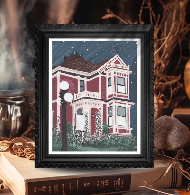 Puede incluir: Una impresi&oacute;n enmarcada de una casa victoriana de noche. La casa est&aacute; pintada en tonos de rojo y blanco, con un cielo azul oscuro salpicado de estrellas. La impresi&oacute;n est&aacute; en un marco negro adornado, apoyada en una pila de libros.