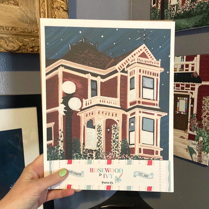 Puede incluir: Una impresi&oacute;n pintada a mano de una casa victoriana con un exterior burdeos y un cielo nocturno azul oscuro. La impresi&oacute;n incluye el texto "Rosewood & Ivy Paper Co. EST 2013". La obra de arte presenta una ilustraci&oacute;n detallada de la casa con molduras blancas y un porche decorativo.