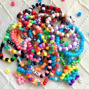 May include: A large collection of colorful plastic pony bead bracelets featuring various solid, translucent, and glitter finishes. Many bracelets include white letter beads spelling words like DAD, ELL, and YEN, alongside scattered loose beads.