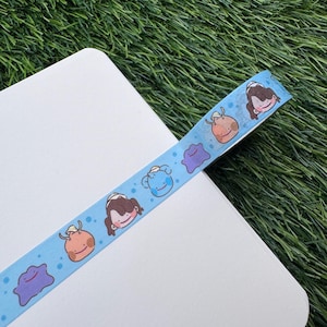 May include: A light blue washi tape featuring cartoon characters and small blue dots. The tape is decorated with various cartoon characters in different colors, including purple, orange, and blue. The tape is placed on a white surface.