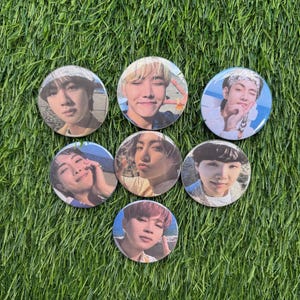 May include: A collection of seven round pinback buttons featuring portraits of a musical group. Each button displays a different member, with various hair colors and expressions. The buttons are arranged on a bed of green grass.