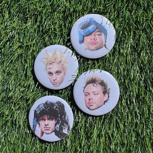 Può includere: Quattro bottoni rotondi bianchi con immagini di volti. I volti hanno acconciature ed espressioni diverse. Un volto indossa un guanto blu e bianco. Lo sfondo è erba verde.