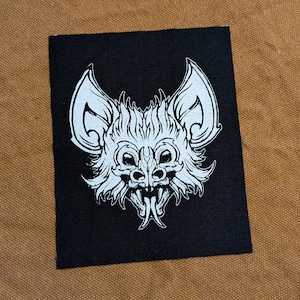 May include: Black and white embroidered patch of a bat with a snarling expression. The bat has large ears and sharp teeth.