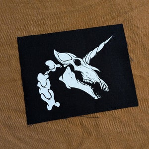 May include: Black fabric patch with a white silhouette of a unicorn skull with a horn. The unicorn skull is facing right and has an open mouth with teeth.