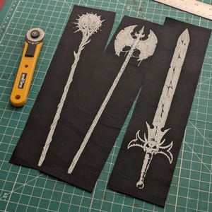 Puede incluir: Tres diseños de armas de fantasía sobre tela negra. Los diseños incluyen un bastón con una esfera, un hacha de doble filo y una espada con una empuñadura en forma de calavera. Un cortador rotatorio amarillo y una regla están en el fondo.