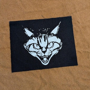 May include: Black fabric patch with a white image of a cat's face with a menacing expression. The cat's eyes are wide open and its mouth is open in a snarl.