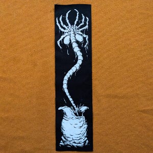 May include: Black and white embroidered patch of an alien creature emerging from a chestburster. The alien has a long, segmented tail and is depicted in a classic horror style.