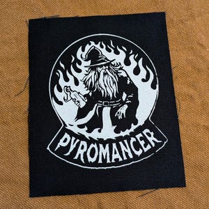 May include: Black and white patch with a wizard holding a lit torch in front of flames. The text "PYROMANCER" is below the wizard.