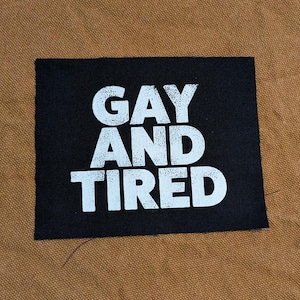 May include: Black fabric patch with white text that reads "Gay and Tired".