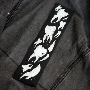 May include: Black rectangular patch featuring a white illustration of teeth in various shapes and sizes. The patch is placed on a dark gray denim jacket, showcasing the contrast between the black and white design and the jacket's texture.