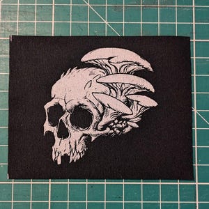 May include: Black and white embroidered patch featuring a skull with mushrooms growing out of the top. The skull is detailed with teeth and eye sockets.