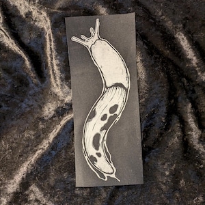 May include: A black and white illustration of a slug on a black background. The slug is depicted in a curled position with a spotted pattern.