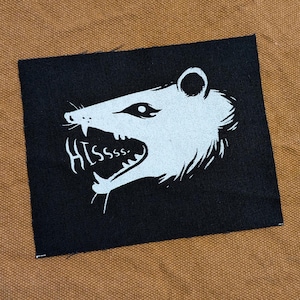 May include: Black fabric patch with a white illustration of an animal with a large mouth and the word "HISSSS." printed below it.
