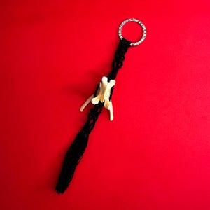 Puede incluir: Un llavero de macramé negro con un anillo plateado y un dije de hueso. El dije de hueso es blanquecino y tiene una forma única. El llavero está sobre un fondo rojo vibrante.