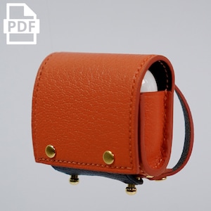 May include: A small, orange leather case with a black strap and gold accents. The case is designed to hold a pair of wireless earbuds.