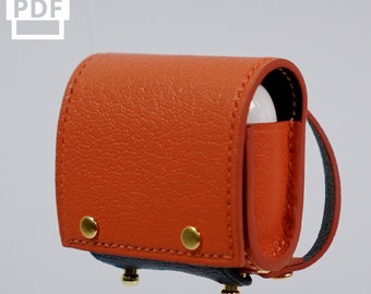 Könnte beinhalten: Eine kleine, orangefarbene Ledertasche mit einem schwarzen Riemen und goldenen Akzenten. Die Tasche ist für ein Paar drahtlose Ohrhörer konzipiert.