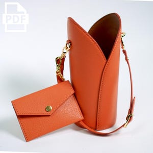 May include: A bright orange leather bucket bag with a gold chain strap and a matching orange leather wallet. The bag has a unique, folded design.