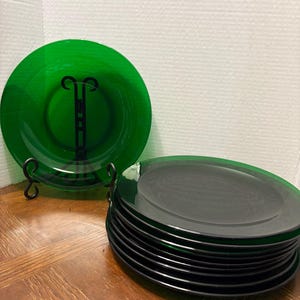 以下が含まれることがあります： 半透明のエメラルドグリーンのガラス製プレートのセット。1枚のプレートが黒い金属製のスタンドに立てかけられ、中央に黒い装飾模様が見えます。その横には、同じ緑色のガラス製プレートが積み重ねられています。