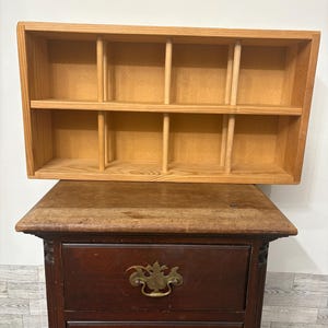 May include: A rectangular light-colored wooden display shelf featuring two horizontal tiers divided by vertical dowels, creating ten individual storage compartments. The unit has a natural wood grain finish and an open front design.