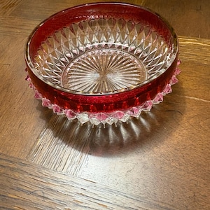 Può includere: Una ciotola di vetro trasparente con un bordo rosso rubino e un motivo decorativo strutturato. La ciotola ha un disegno a stella sul fondo e un bordo smerlato. La ciotola è probabilmente per servire o esporre.