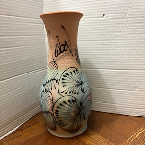 May include: A terracotta-coloured ceramic vase with a flared rim and a narrow neck. The lower body features a gradient of soft blue and pink hues, decorated with black hand-painted fan-shaped floral patterns. The text "Abad" is written in black.