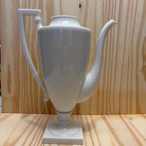 Może przedstawiać: Biały ceramiczny czajnik o unikalnym, kanciastym designie. Czajnik ma wysoki, cylindryczny korpus, zakrzywioną wylewkę i kwadratową podstawę. Uchwyt ma prostokątny kształt. Czajnik jest wystawiony na drewnianej powierzchni.