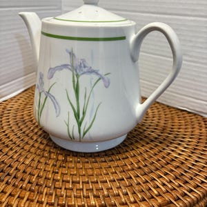 May include: A white porcelain teapot featuring a delicate floral pattern of light purple iris flowers with green stems and leaves. The teapot has a matching lid with a finial, a curved handle, a spout, and a thin green band encircling the top rim.