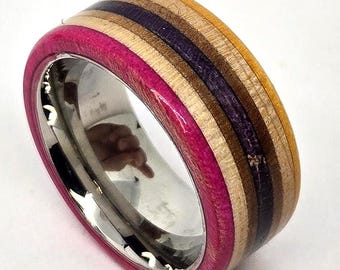 May include: A ring featuring a polished silver-toned metal inner band and an outer band constructed from multiple layered materials. The exterior displays horizontal stripes of vibrant pink, light tan, dark brown, deep purple, and golden yellow.