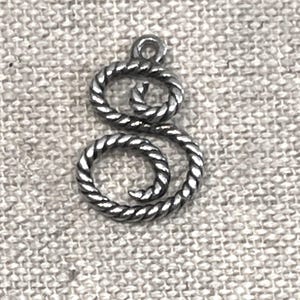 May include: A silver-colored metal charm in the shape of a stylized letter "S". The charm has a twisted rope design and a small loop at the top for attaching to a necklace or bracelet. The charm is shown on a textured, light gray background.