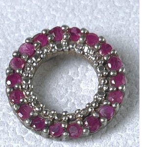 May include: A circular pendant featuring a double row of small, round, deep pink rubies set in a silver-toned metal frame. The design consists of two concentric circles of gemstones separated by a thin band of textured, beaded silver-toned metal.