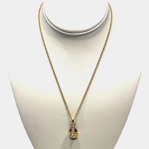 May include: A gold-colored necklace with a pendant shaped like an egg. The pendant has a red base, gold-colored accents, and small, sparkling gems. The necklace is displayed on a white mannequin.