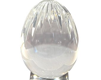 May include: A clear, egg-shaped crystal sculpture with vertical grooves at the top. The sculpture sits on a square, clear crystal base. The overall design is elegant and decorative.