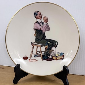 May include: A decorative plate featuring a seated man sewing, with a small dog and sewing supplies. The plate has a gold rim and is displayed on a black stand. The artist's signature, "Norman Rockwell," is visible.