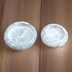 May include: Two round, light gray concrete bowls with a speckled pattern. The bowls have a shallow, concave interior and a smooth, rounded exterior. The bowls are of different sizes and are suitable for holding small items.