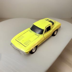 May include: A bright yellow toy car with a black windshield and silver trim. The classic car has a sleek design with visible details like the hood vents and wheels. The toy car is a miniature replica of a vintage sports car.