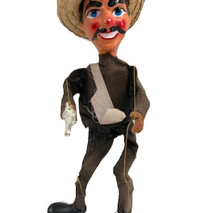 May include: A hand puppet of a man wearing a brown and white straw hat, a brown shirt, black pants, and black shoes. He has a painted face with a mustache and a gun in his hand.