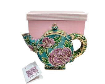 May include: A small, green and pink teapot with floral designs and gold accents. The teapot is sitting on a pink box with white stripes. The box has a label that says "NYCO".